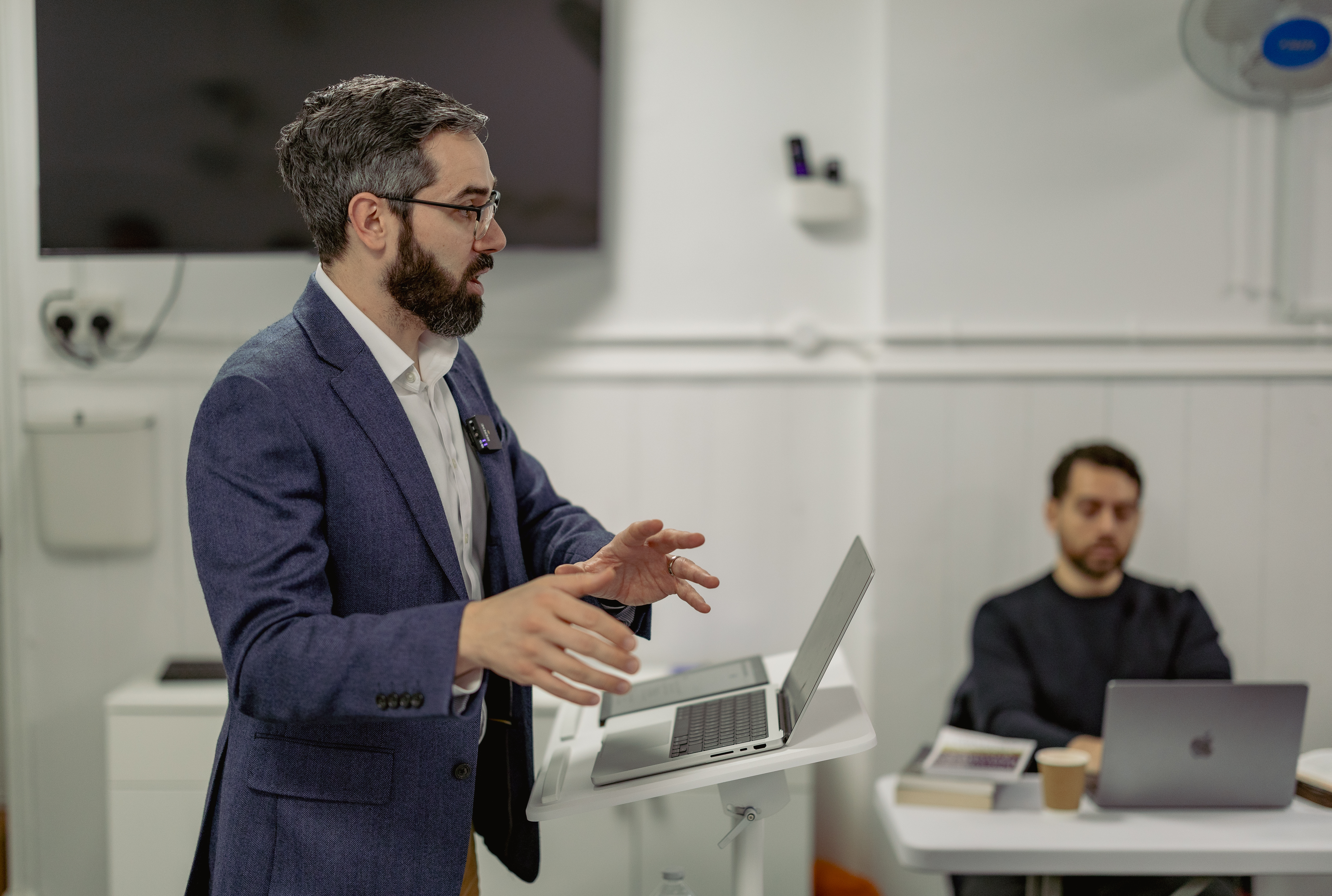 Instructor delivering a lecture for the Hermeneia Diploma in Expository Preaching, speaking at a podium with a laptop while a student listens in a classroom setting.